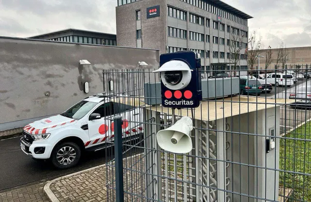 Eine Securitas-Kameraeinheit mit Lautsprecher ist an einem Zaun befestigt, im Hintergrund ein Securitas-Firmengebäude und ein Einsatzfahrzeug auf dem Betriebsgelände.