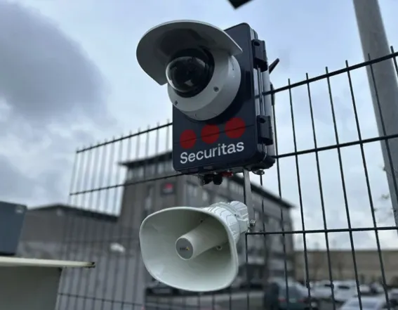 SBC-Kameraeinheit in Nahaufnahme mit Lautsprecher Nahaufnahme einer Securitas SBC-Einheit mit Kamera und Lautsprecher, montiert an einem Maschendrahtzaun vor einem Bürokomplex.