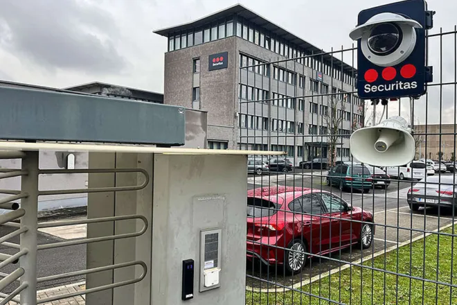 Zugangsschleuse mit SBC-Kamera am Werkseingang Eine Zutrittskontrollschleuse mit Lesegerät und Securitas SBC-Kameraeinheit am Zaun, im Hintergrund das Securitas-Bürogebäude.